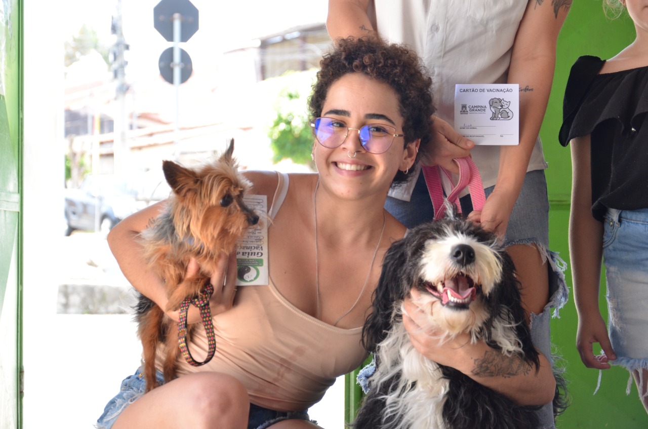 Mulher sorrindo abraçando cachorros e com cartão de vacina na mão